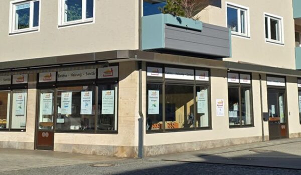 Eckansicht des Firmensitzes der Schröder GmbH. Das Gebäude ist modern und beige mit großen Fenstern, auf denen Schilder zu sehen sind. Im Erdgeschoss befindet sich die Schröder GmbH mit Plakaten an den Fenstern. Die oberen Stockwerke haben Balkone mit Pflanzen. Die Straße ist mit Steinen gepflastert.