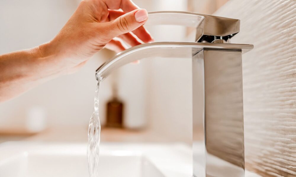 Eine Nahaufnahme der Hand einer Person, die einen eleganten, modernen Chromhahn mit gebogenem Ausguss aufdreht. Wasser fließt in ein weißes Becken darunter. Der Hintergrund ist leicht verschwommen und lenkt die Aufmerksamkeit auf den Hahn.