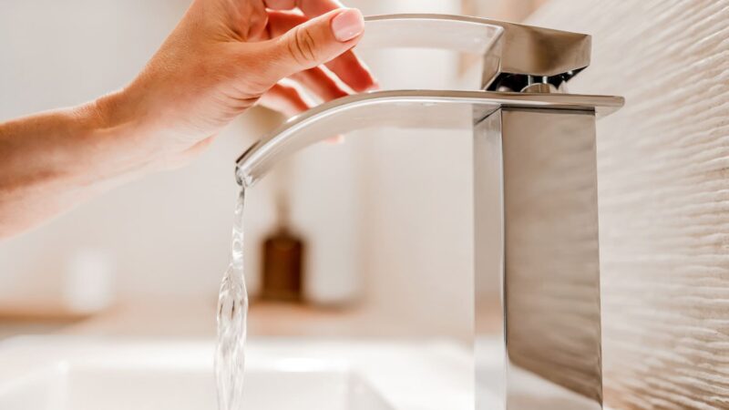 Eine Nahaufnahme der Hand einer Person, die einen eleganten, modernen Chromhahn mit gebogenem Ausguss aufdreht. Wasser fließt in ein weißes Becken darunter. Der Hintergrund ist leicht verschwommen und lenkt die Aufmerksamkeit auf den Hahn.