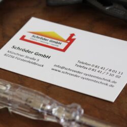 Eine Visitenkarte der Schröder GmbH, die zwischen verschiedenen Werkzeugen auf dem Tisch liegt, zeigt Kontaktinformationen wie eine Telefonnummer, eine E-Mail-Adresse und die Postanschrift in Fürstenfeldbruck.
