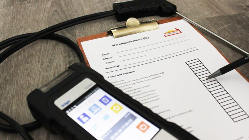 Eine Wartungscheckliste auf einem Klemmbrett liegt auf einer Holzoberfläche. Ein Stift zeigt auf Kontrollkästchen auf dem Papier. Neben dem Klemmbrett liegt ein schwarzes elektronisches Gerät mit einem digitalen Bildschirm und verschiedenen Symbolen.