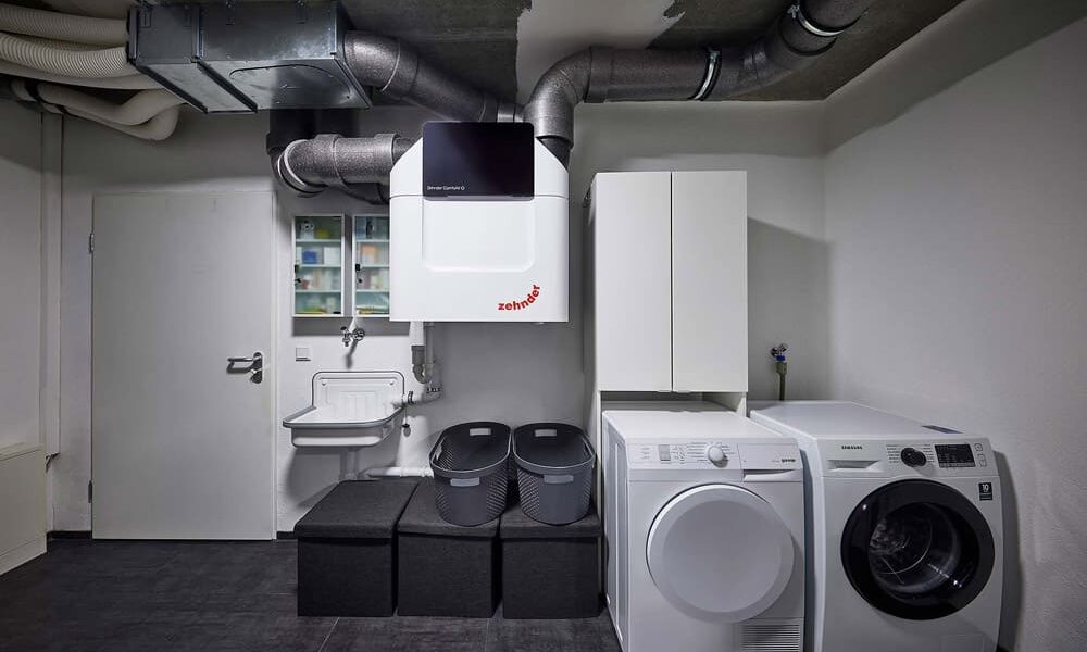 Ein moderner Waschraum mit Waschmaschine und Trockner nebeneinander, Belüftungssystem darüber, Lagerschränken, einem wandmontierten Waschbecken und drei Lagerbehältern. Der Raum hat einen dunklen Boden und helle Wände.