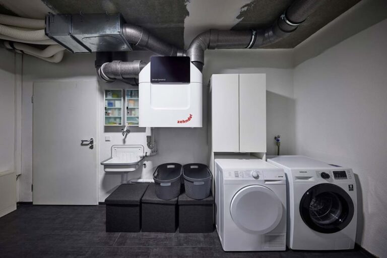 Ein moderner Waschraum mit Waschmaschine und Trockner nebeneinander, Belüftungssystem darüber, Lagerschränken, einem wandmontierten Waschbecken und drei Lagerbehältern. Der Raum hat einen dunklen Boden und helle Wände.