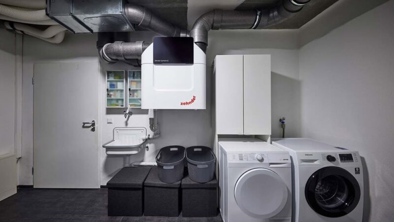 Ein moderner Waschraum mit Waschmaschine und Trockner nebeneinander, Belüftungssystem darüber, Lagerschränken, einem wandmontierten Waschbecken und drei Lagerbehältern. Der Raum hat einen dunklen Boden und helle Wände.